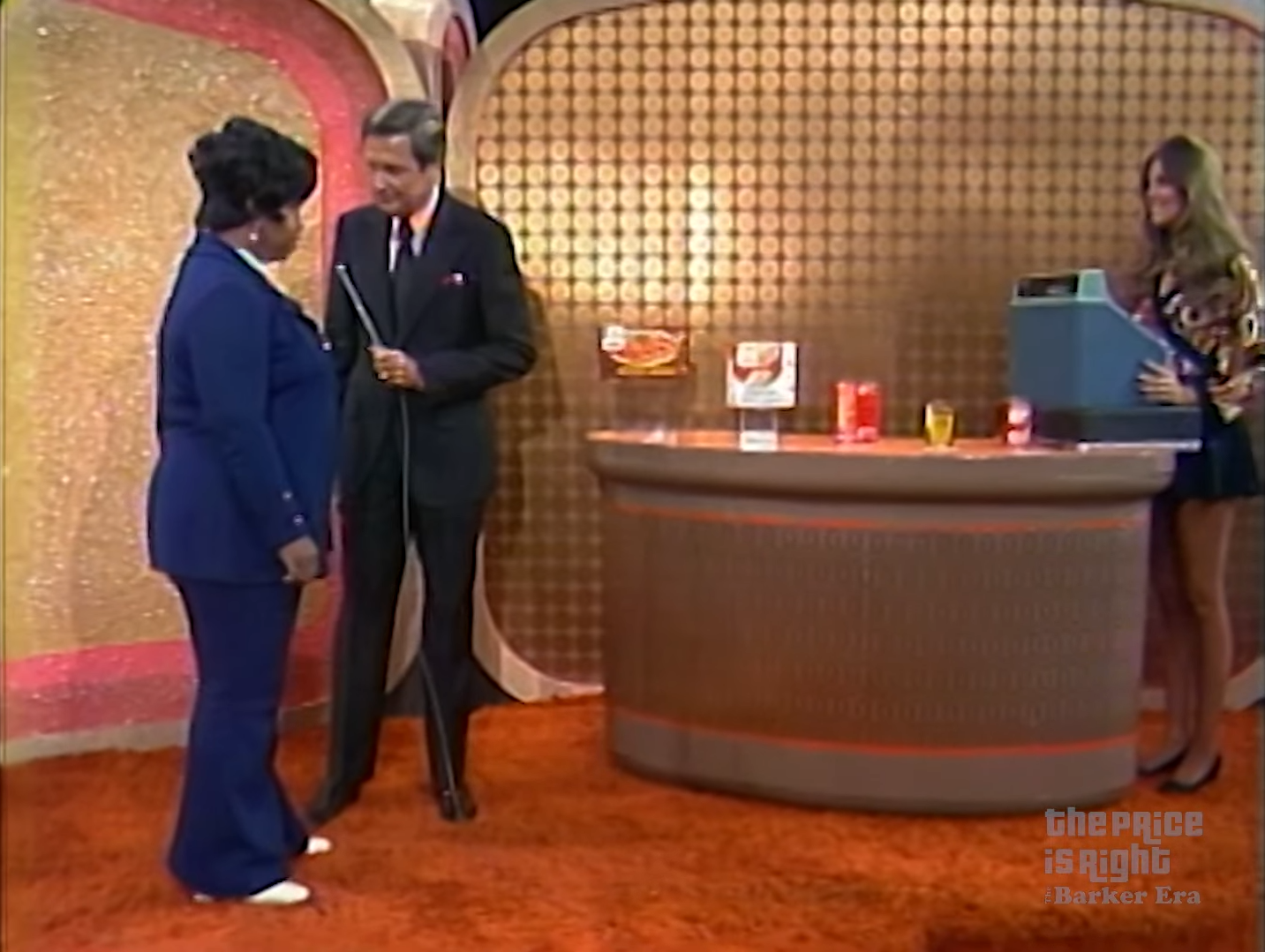 A contestant on The Price Is Right stands speaking to the host while a model prepares to ring up groceries on a cash register.