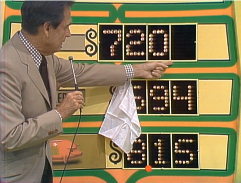 Host pointing at the pricing board for the game 'Any Number', showing three prices: 0, 4, and 5, with one digit missing from each. A microphone is in the host's hand, and a white cloth is draped over it.