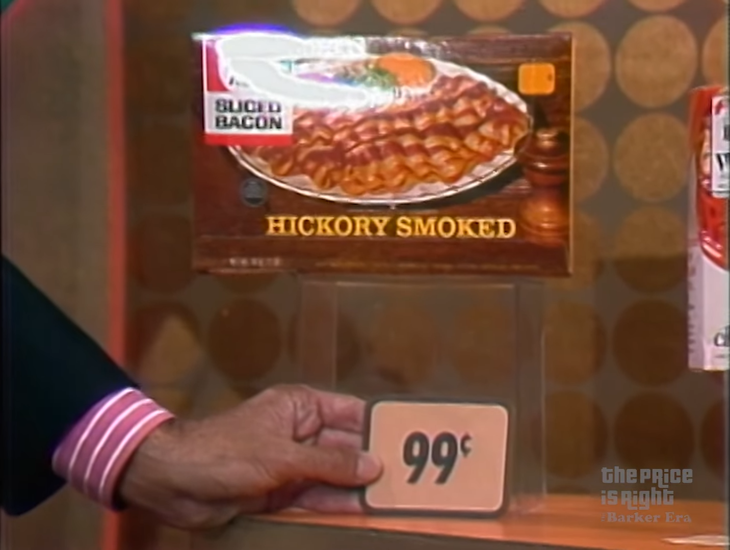 A hand holding a price tag of 99 cents in front of a packaging for hickory smoked sliced bacon on a display.