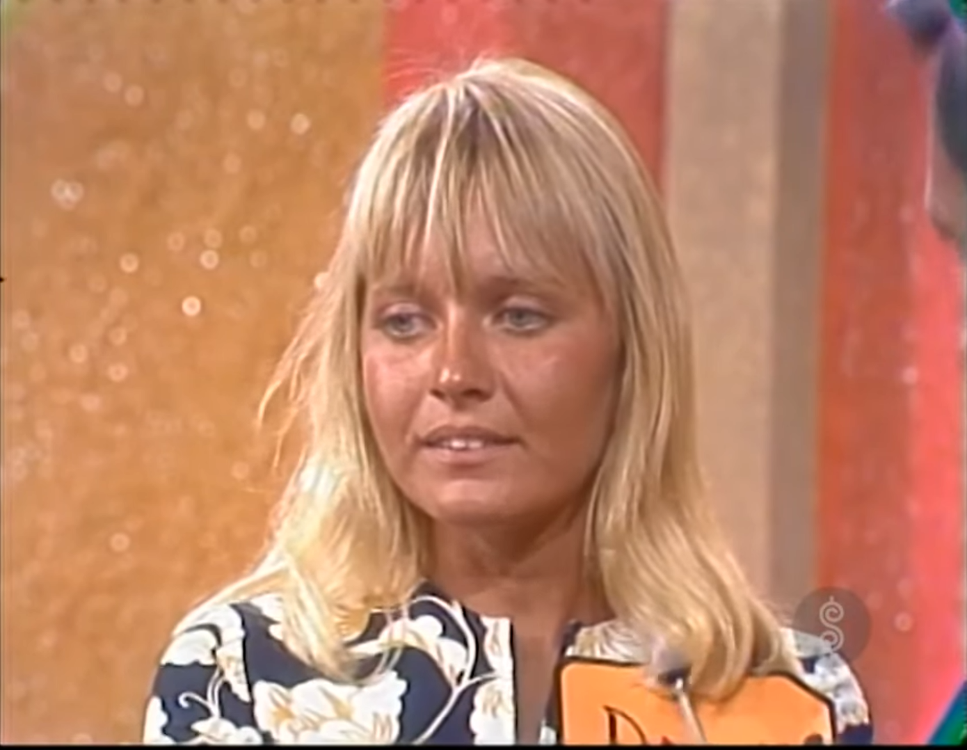 Close-up of a contestant with blonde hair, wearing a floral patterned blouse, looking intently while holding a bid card on a game show set.