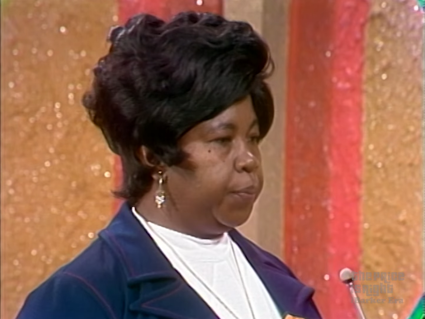 A contestant listening intently during the Grocery Game on The Price is Right, wearing a blue jacket and earrings, with a colorful backdrop.