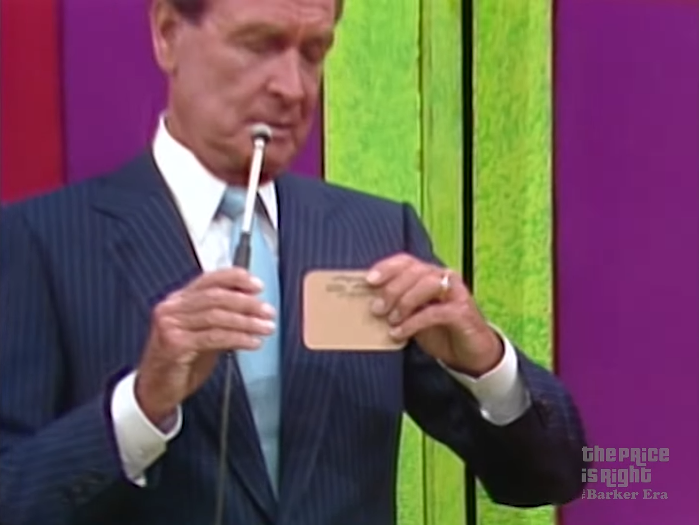 A man in a suit holds a card and microphone while standing in front of a colorful backdrop on a game show set.
