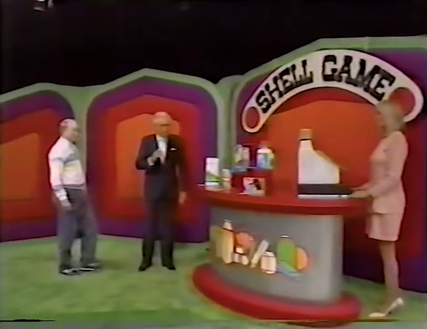 A game show scene with a vibrant background featuring the title 'SHELL GAME'. A male contestant stands next to the host, while a female model operates a cash register displaying various grocery items.