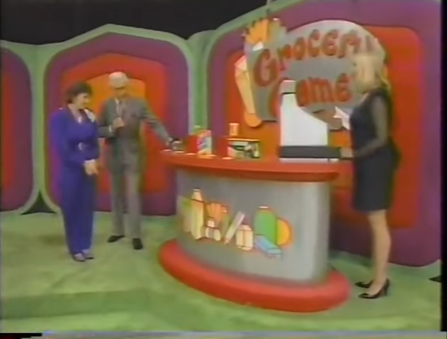 A contestant stands next to a host at a colorful set for the Grocery Game, with grocery products displayed on a counter. A model operates an old cash register in the background.
