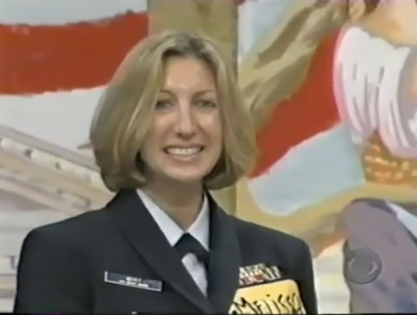 A smiling contestant wearing a navy blue jacket with a name tag and a yellow contestant sign, standing in front of a colorful background.