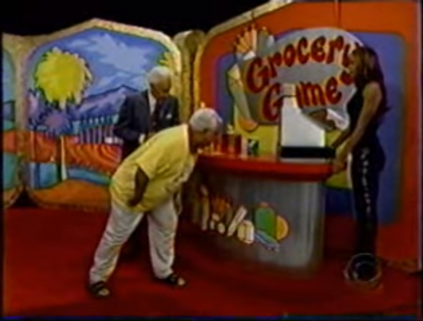 A contestant in a yellow shirt bends over to examine grocery items while a host stands nearby, with a woman operating an old-fashioned cash register at the Grocery Game set.