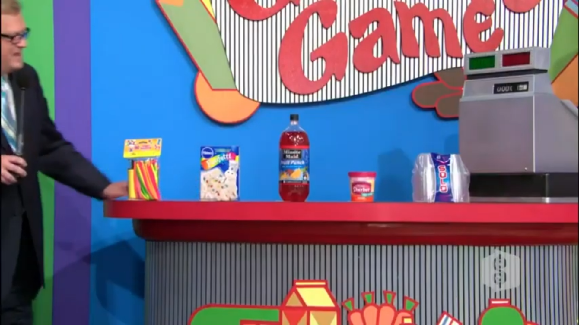 A colorful display of grocery items on a game show set, featuring a bottles of fruit punch, boxes of snack foods, and a cash register.