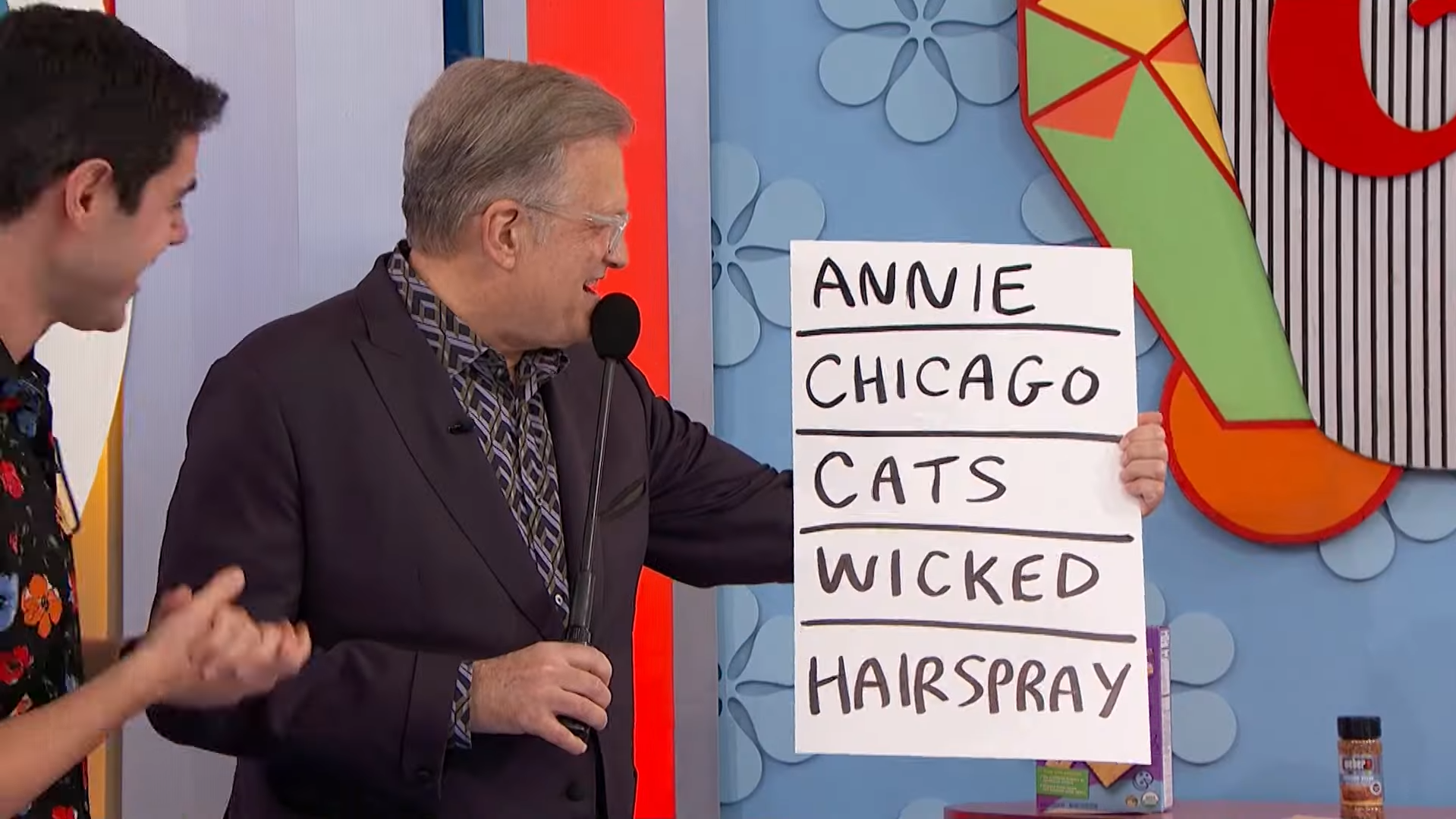 Drew Carey holds a large sign displaying the names of musicals: 'ANNIE', 'CHICAGO', 'CATS', 'WICKED', and 'HAIRSPRAY', while a contestant stands beside him, smiling.