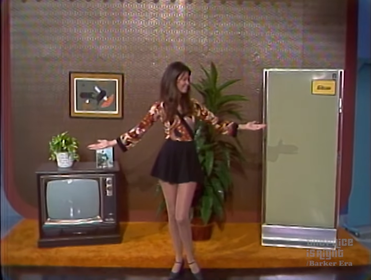 A game show model presents prizes, including a vintage television and a refrigerator, in a colorful studio set.