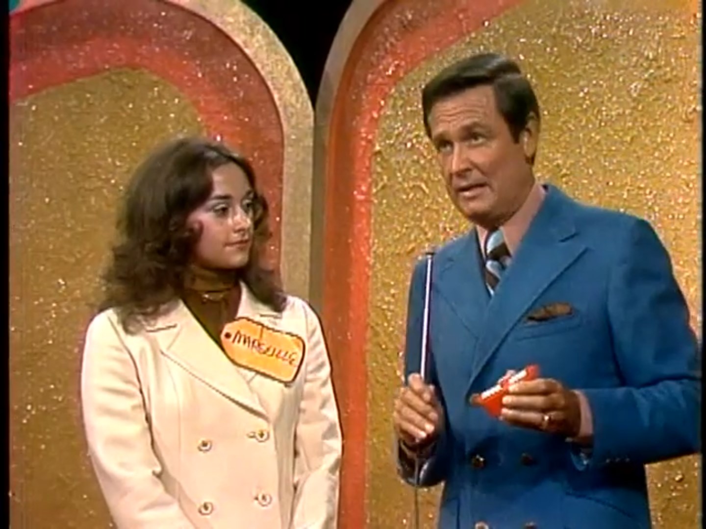 A contestant named Marcella stands next to the host, Bob, during the Clock Game segment. Bob holds a small red object while explaining the game.