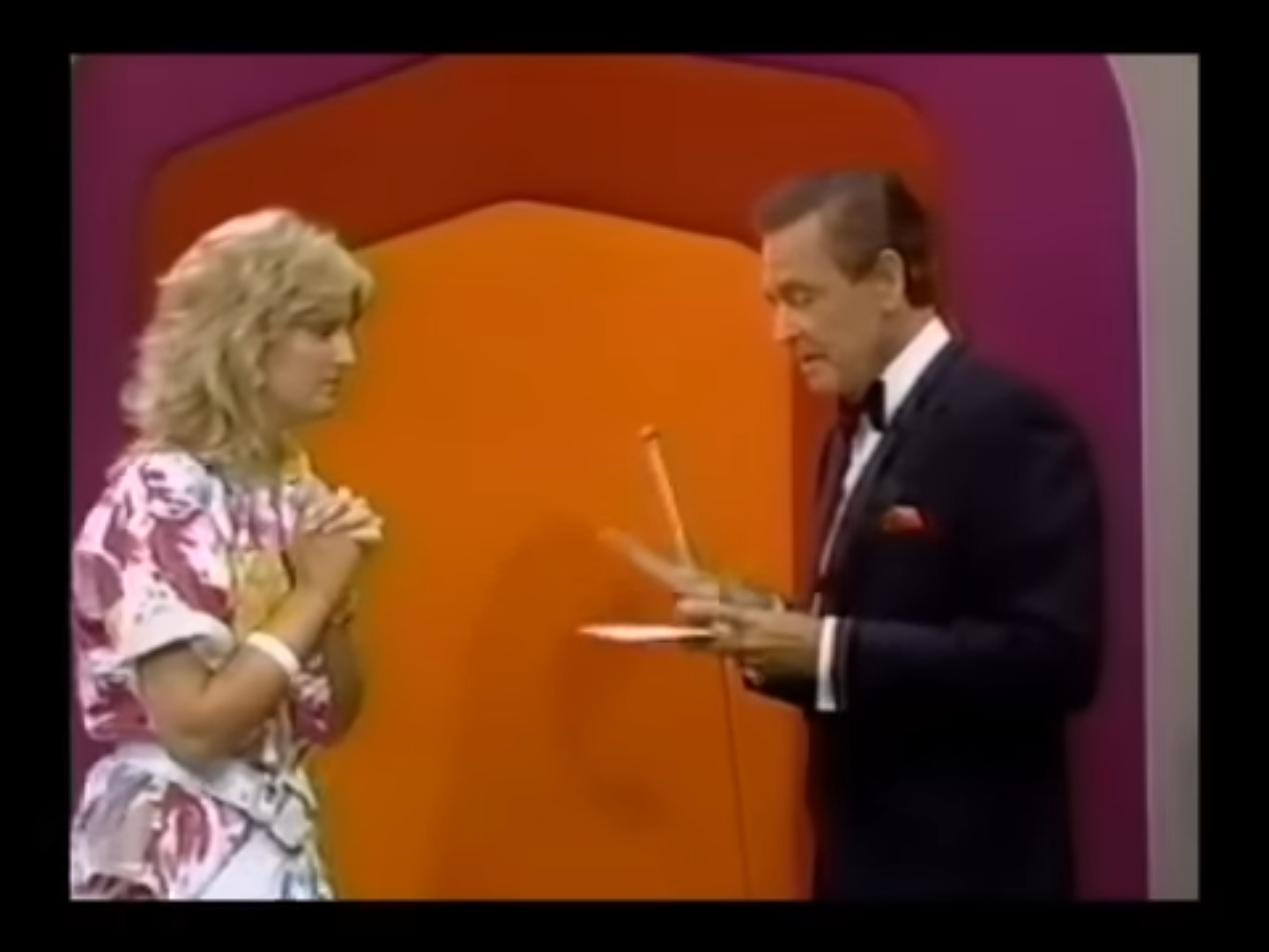 A female contestant nervously stands with her hands clasped in front of her, while the host holds a price card, set against a colorful game show backdrop.