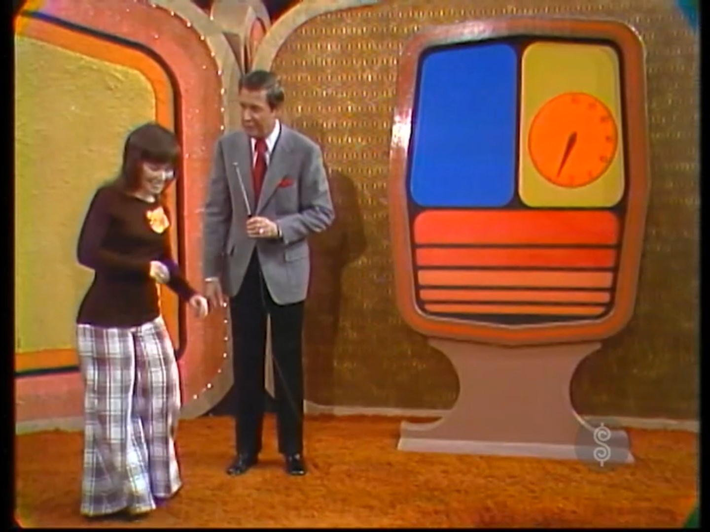 A contestant smiles excitedly while standing next to a game host in a brightly colored studio, with a large game board featuring a clock design behind them.