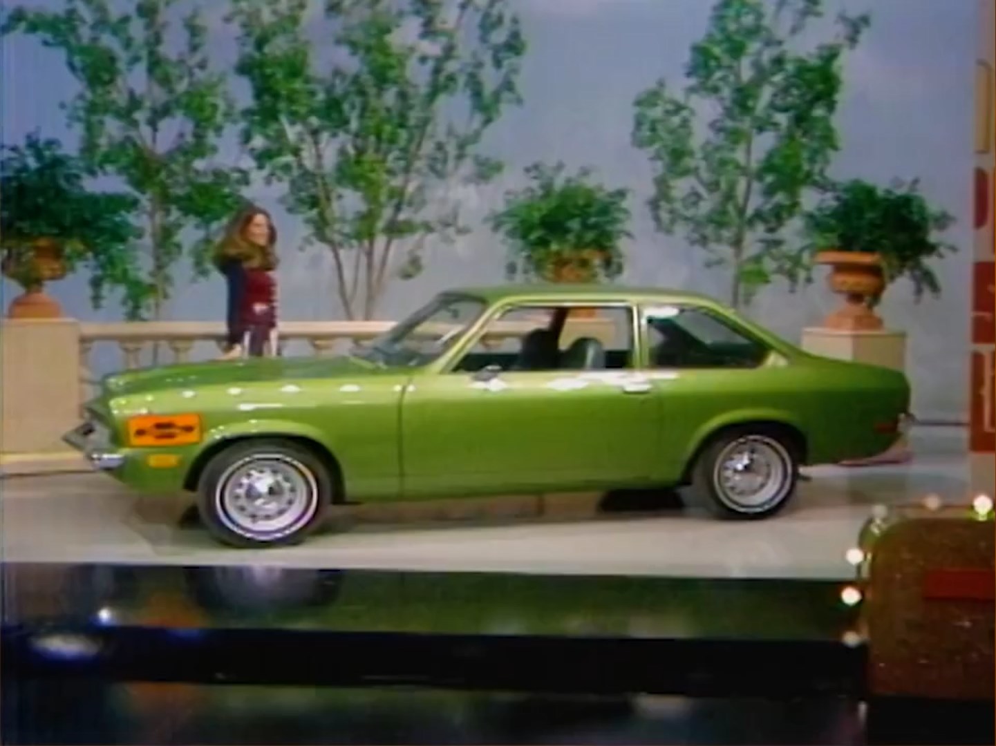 A green Chevy Vega sedan displayed on a game show stage, with a woman in a colorful outfit standing beside it.