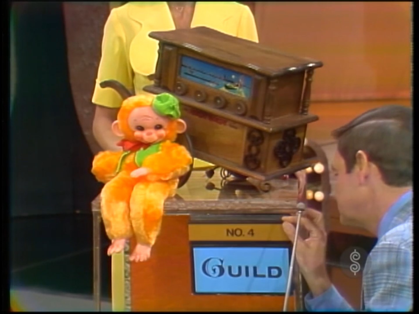 A brightly colored monkey toy sits on a podium beside a vintage wooden music box. A host leans in, speaking to the audience.