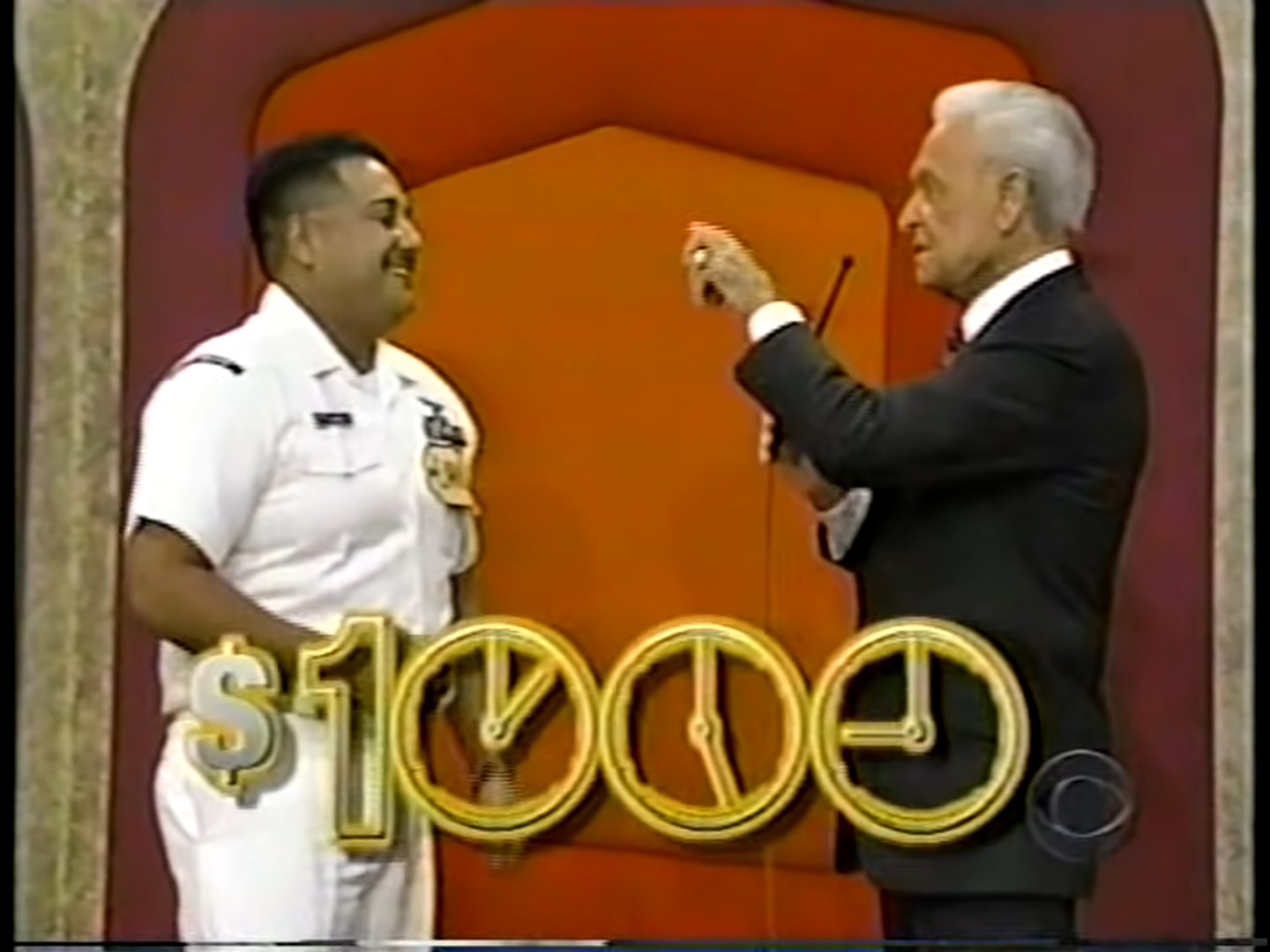 Contestant smiling in a white uniform stands next to the host, holding a microphone, with the prize amount of ,000 displayed prominently.
