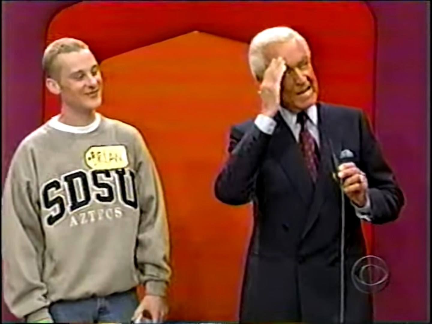 A contestant smiling in a gray sweatshirt stands next to a flabbergasted game show host, who gestures while speaking, in front of a colorful game show backdrop.