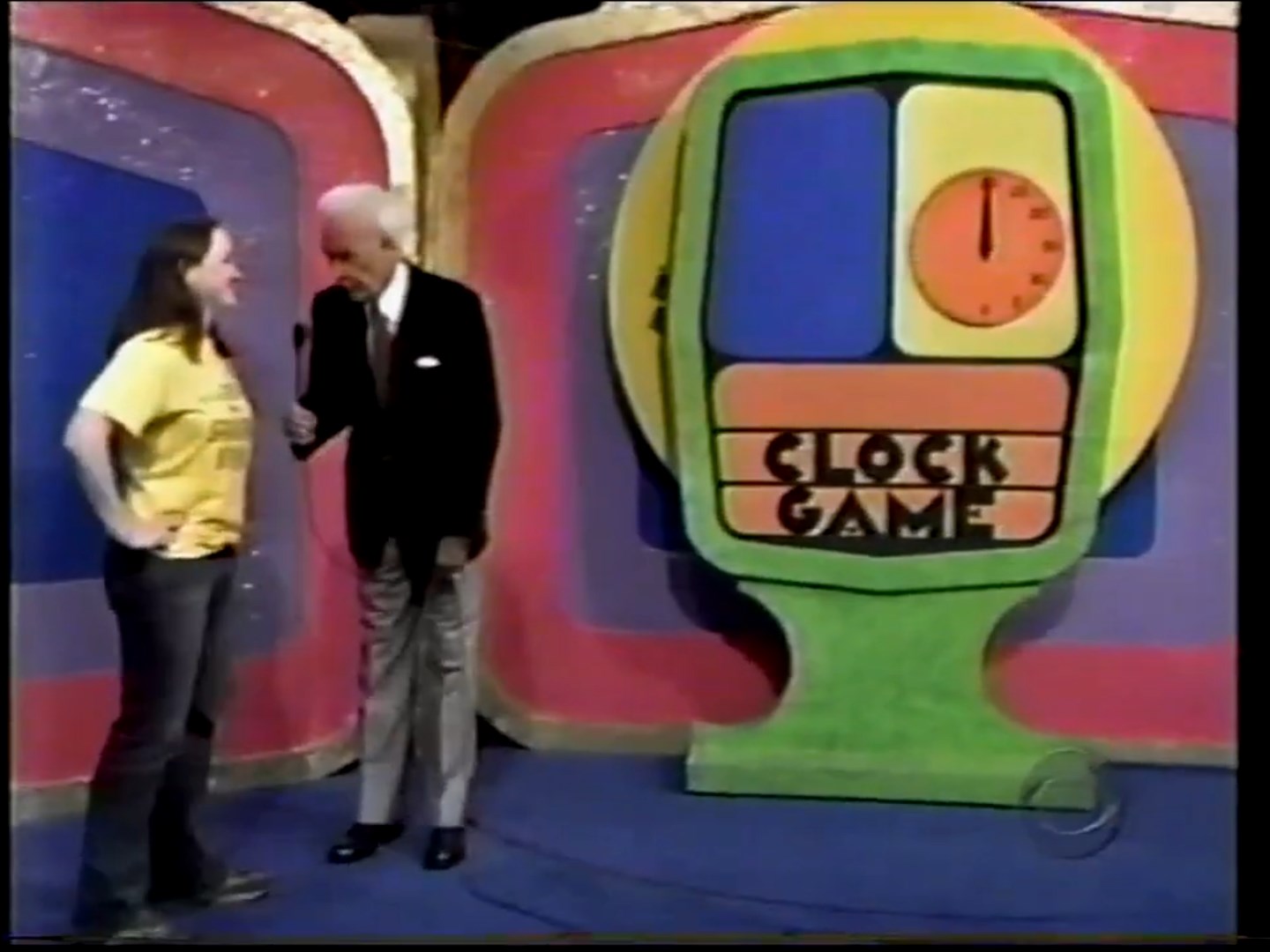 A contestant and host engaged in the Clock Game segment, with a colorful game board featuring a clock design in the background.