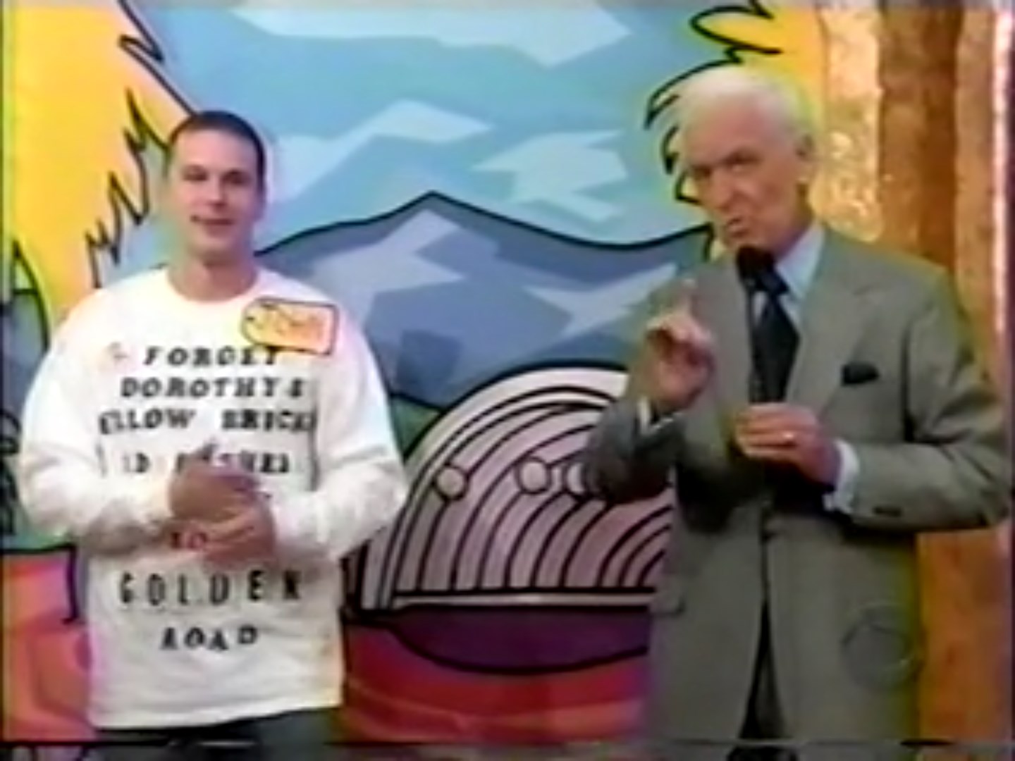 A contestant named John stands next to the host Bob Barker, both smiling and engaged in the Clock Game segment, with a colorful backdrop featuring palm trees and mountains.
