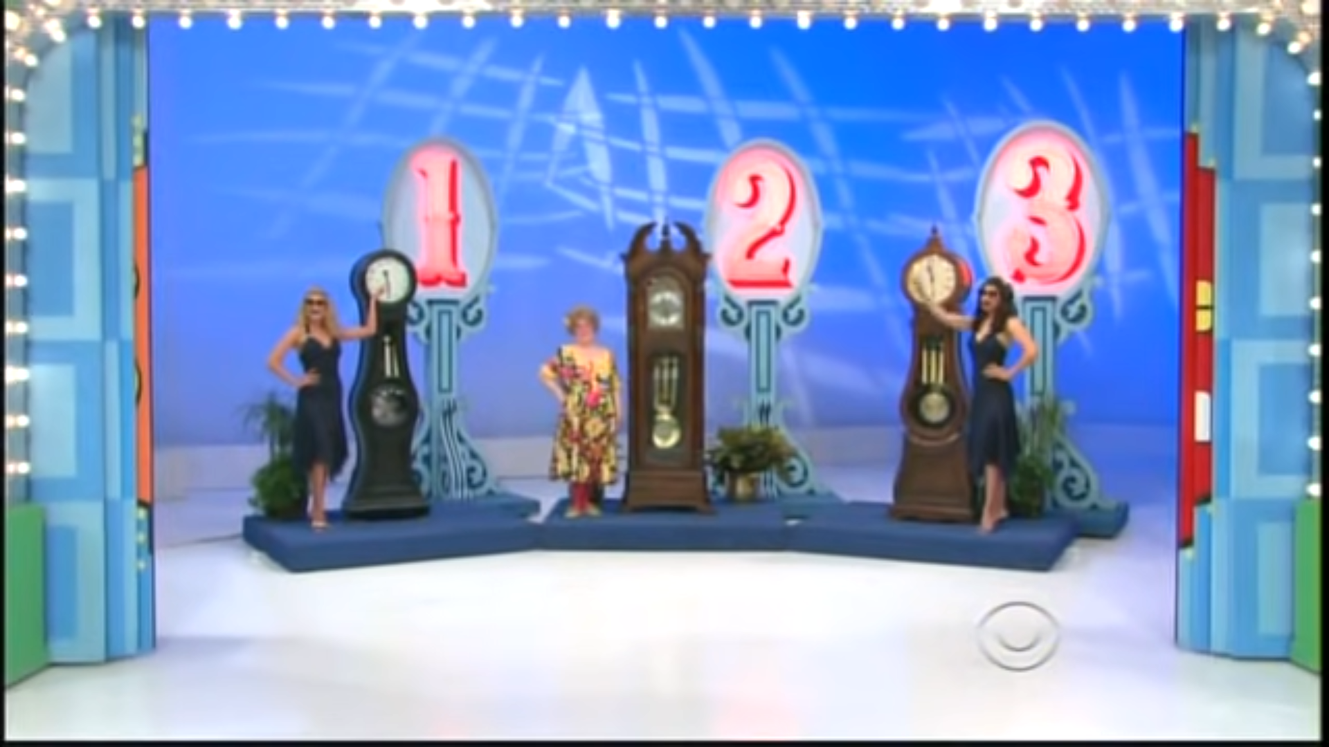 Contestants stand beside three grandfather clocks labeled 1, 2, and 3, as part of the 'Most Expensive' game on TPIR.