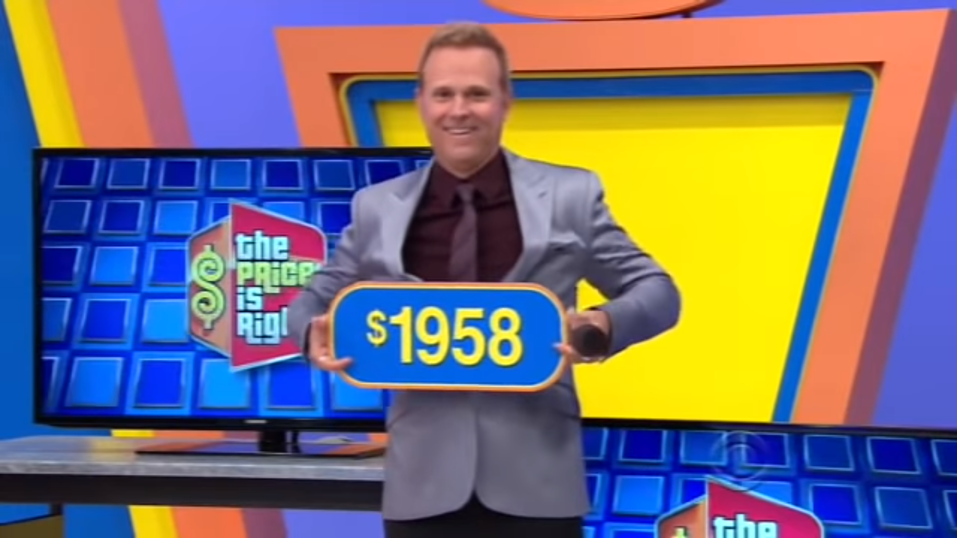 George Gray The Price Is Right smiles while holding a sign displaying the number 58, set against a colorful game show backdrop.