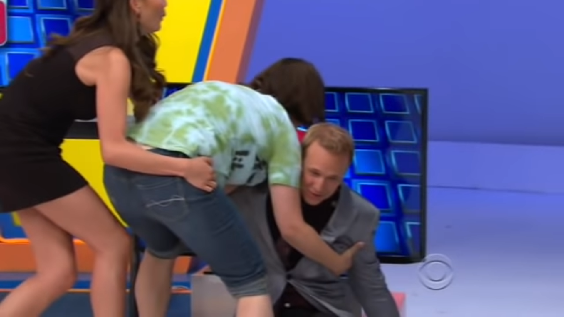 A male contestant is being helped by two women on stage, one appears to be assisting him while he appears to lose balance, creating a moment of excitement during a game show.