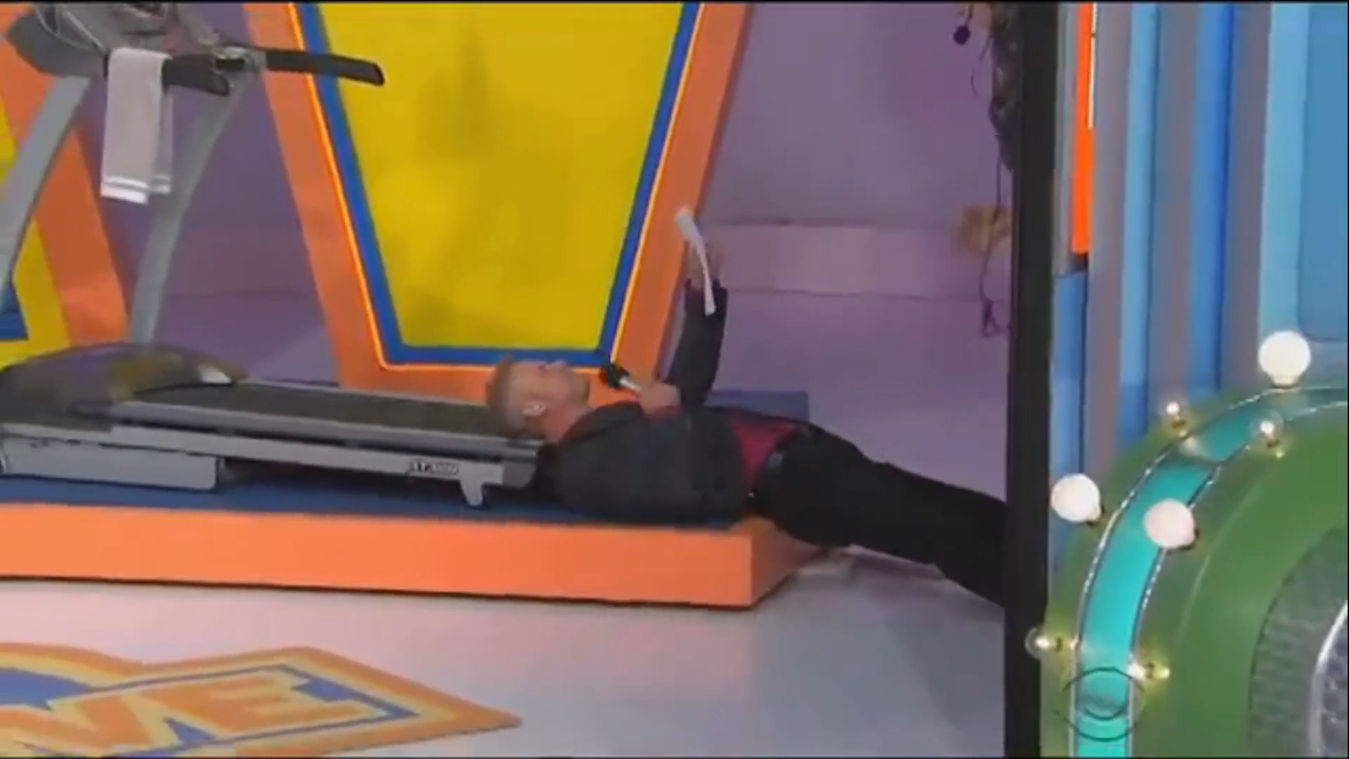 George Gray lies on the ground next to a treadmill, holding a microphone and looking amused.