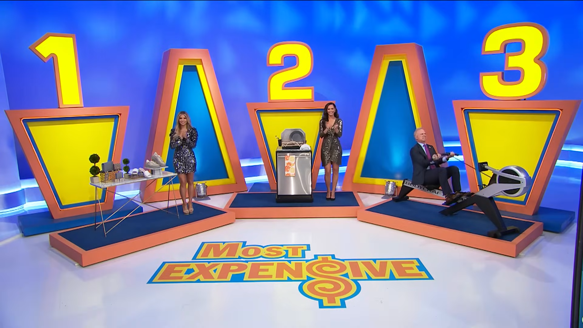 Models standing in front of three prize displays, labeled #1, #2, and #3, in the game show 'Most Expensive'.