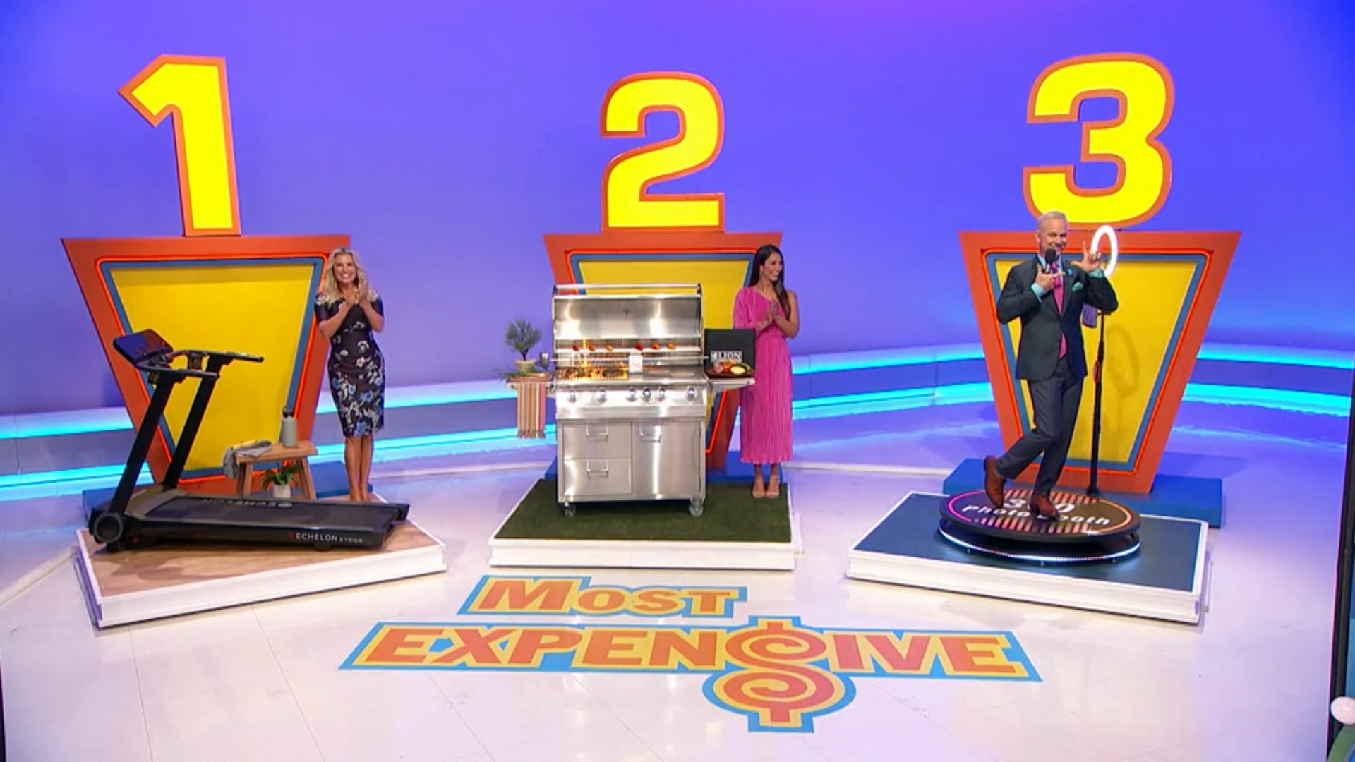 Three prize displays for the game 'Most Expensive', featuring a treadmill for prize #1, a stainless steel grill for prize #2, and a podium with a contestant for prize #3. The backdrop includes large numbers indicating the prizes' order.