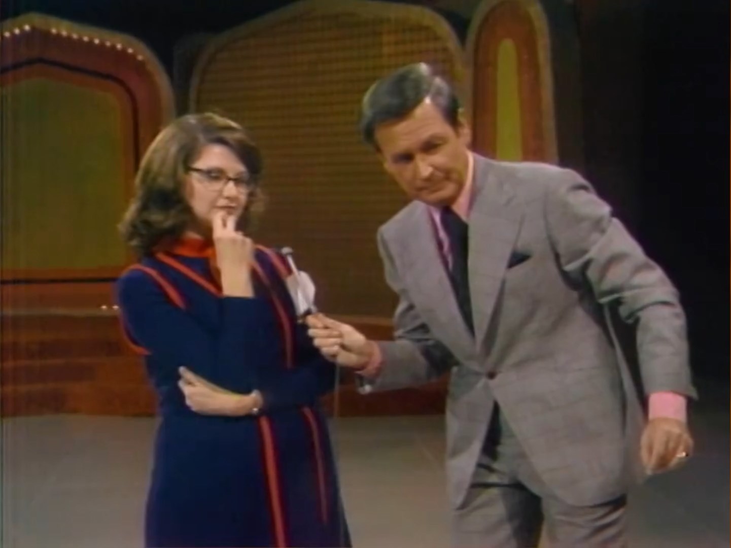 A contestant deep in thought, holding a finger to her mouth, while a game show host gestures towards her during a classic car game on a brightly colored set.