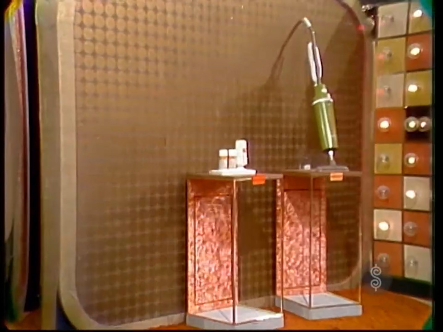 Image of a game show stage setup featuring two small prize displays: one with a green vacuum cleaner and the other with a facial kit. The background includes a patterned wall and a display board with lights.