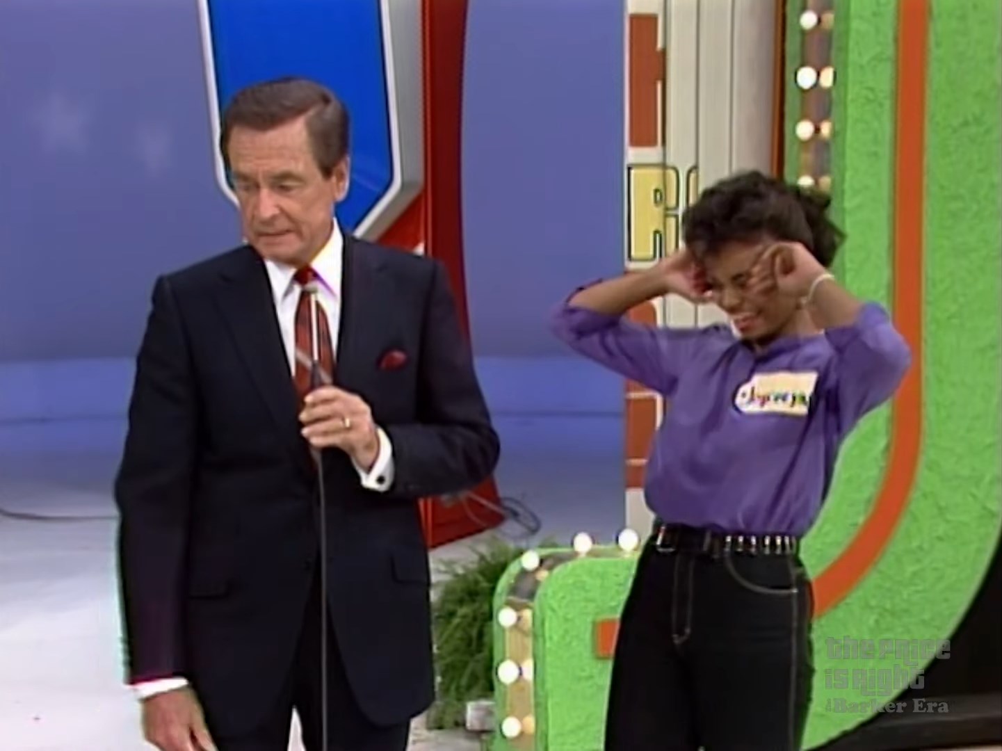 Bob is speaking into a microphone while a contestant in a purple shirt covers her ears in anticipation.