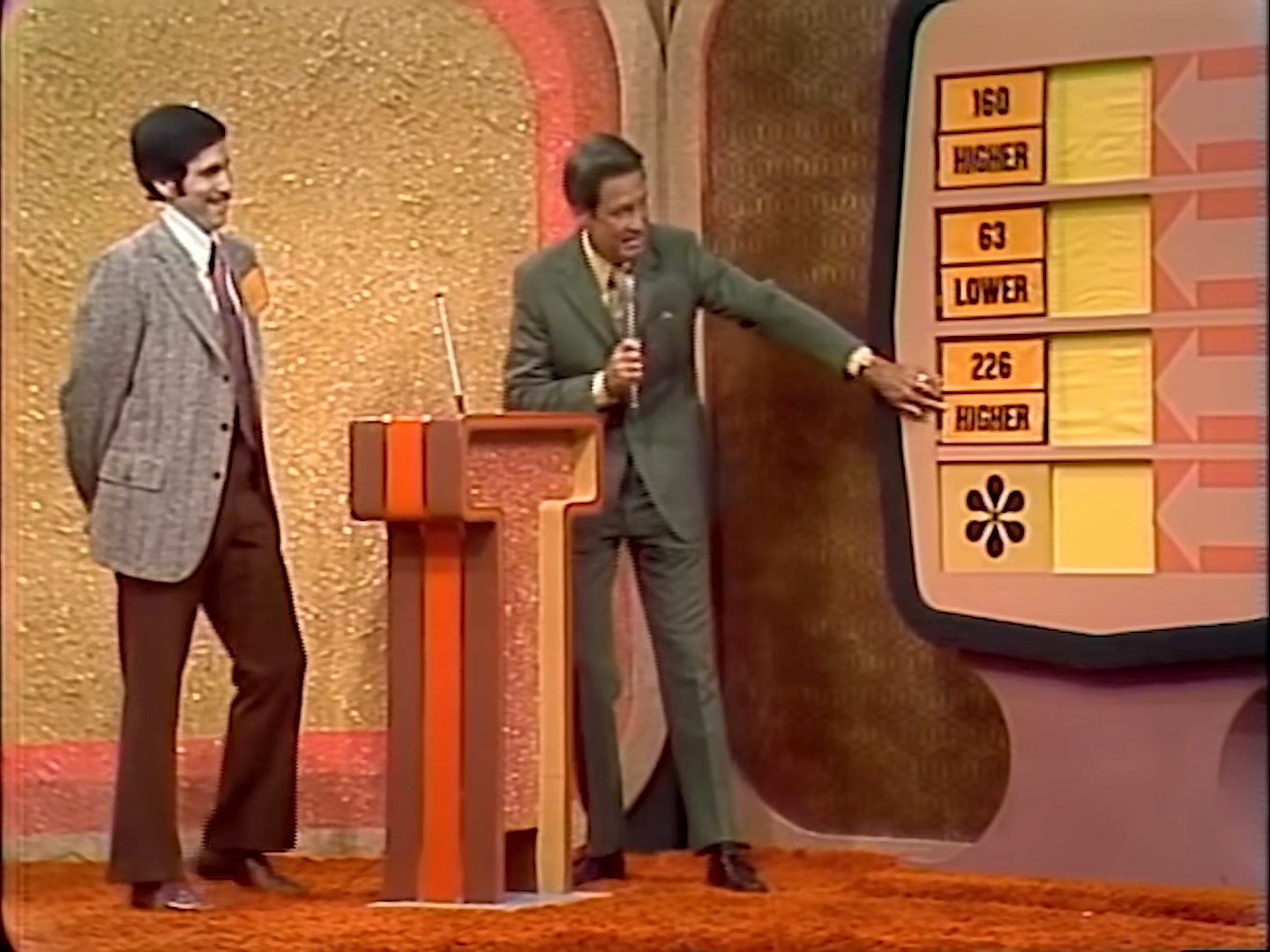 A game show host points to a board displaying numbers while a participant looks on, both smiling during a segment of the Bonus Game.