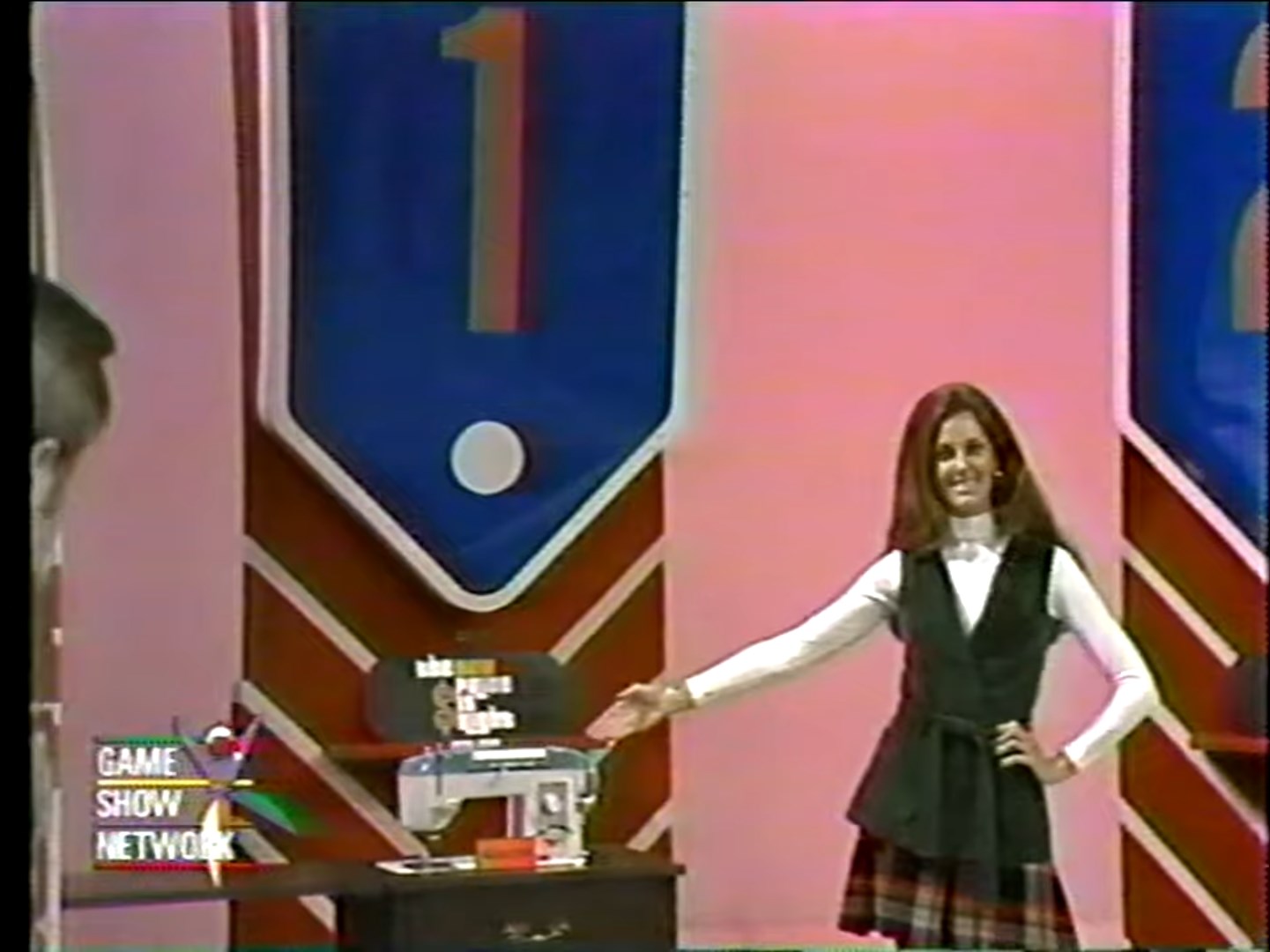 A game show model presents a sewing machine, standing next to a large sign labeled '1'.