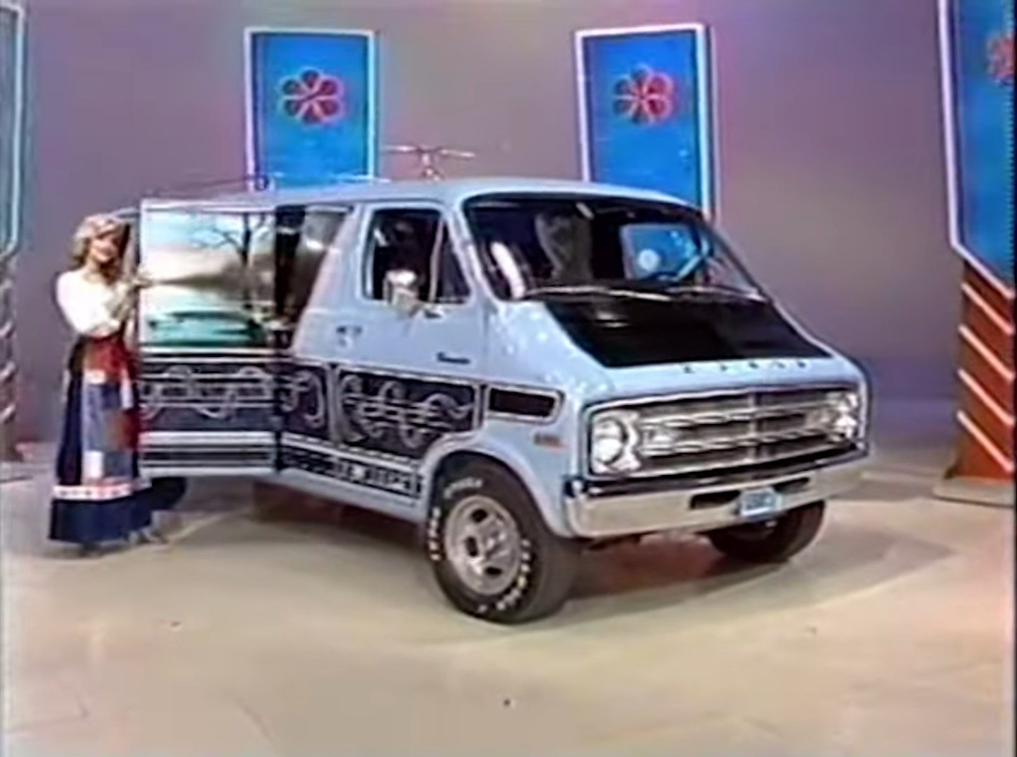 A model showcases a vintage blue van with decorative designs while standing on a game show set.