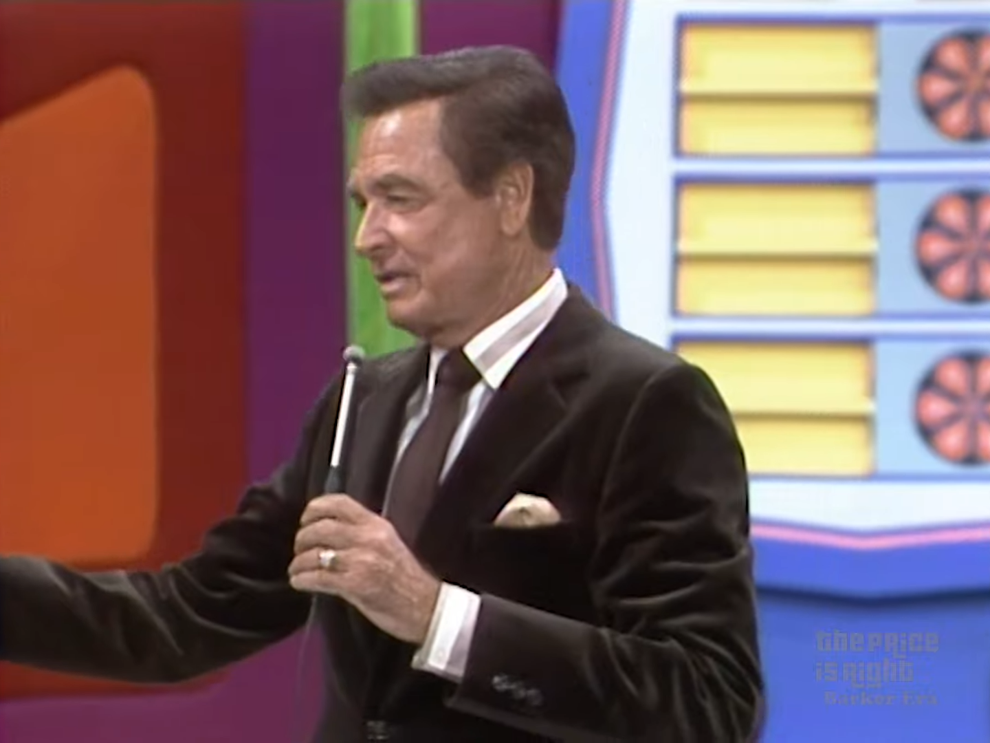 A television game show host speaks into a microphone while gesturing at an unseen contestant, with colorful set decorations in the background.