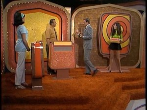 A scene from The Price is Right in 1972 featuring a host playing 'Double Bullseye' with two contestants, with colorful game boards and a vibrant set design.