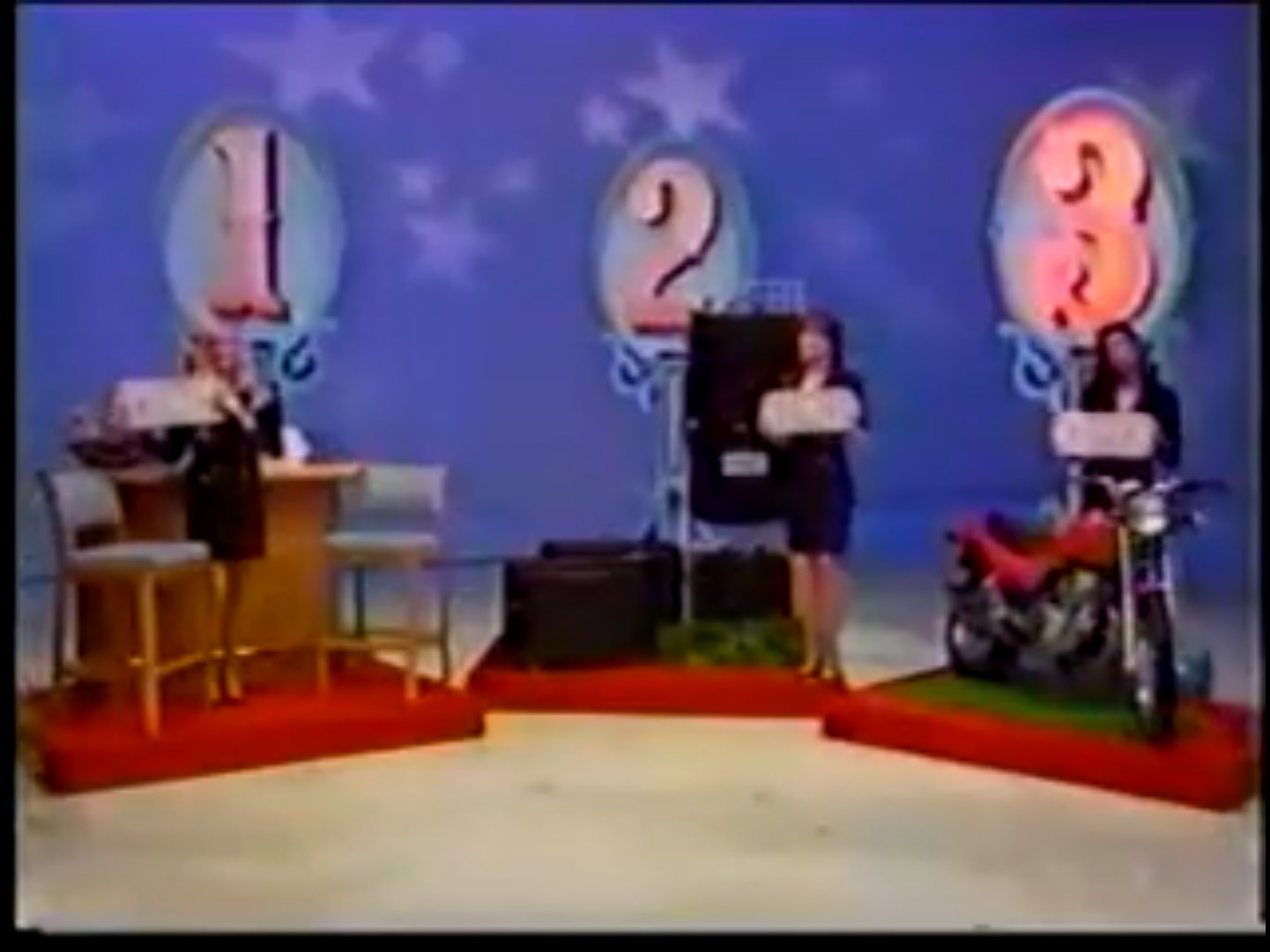 A game show set featuring three podiums labeled 1, 2, and 3, showcasing different prizes, with a host and two contestants.
