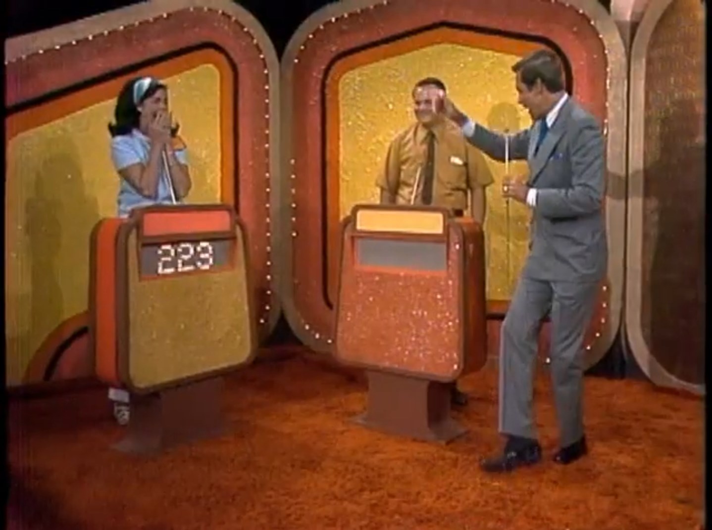 A female contestant excitedly reacts behind her podium while a male contestant smiles next to her. A game show host gestures with a microphone, surrounded by colorful game show set elements.