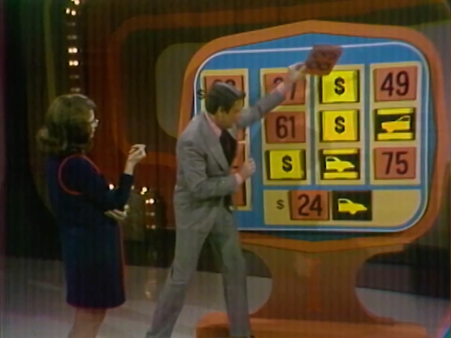A game host reveals a number on a colorful board while a contestant watches, with various numbers and dollar signs visible.