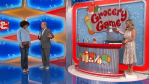 A scene from The Price is Right featuring two contestants and a host interacting near a game called 'Grocery Game,' with a colorful backdrop and items displayed on the counter.