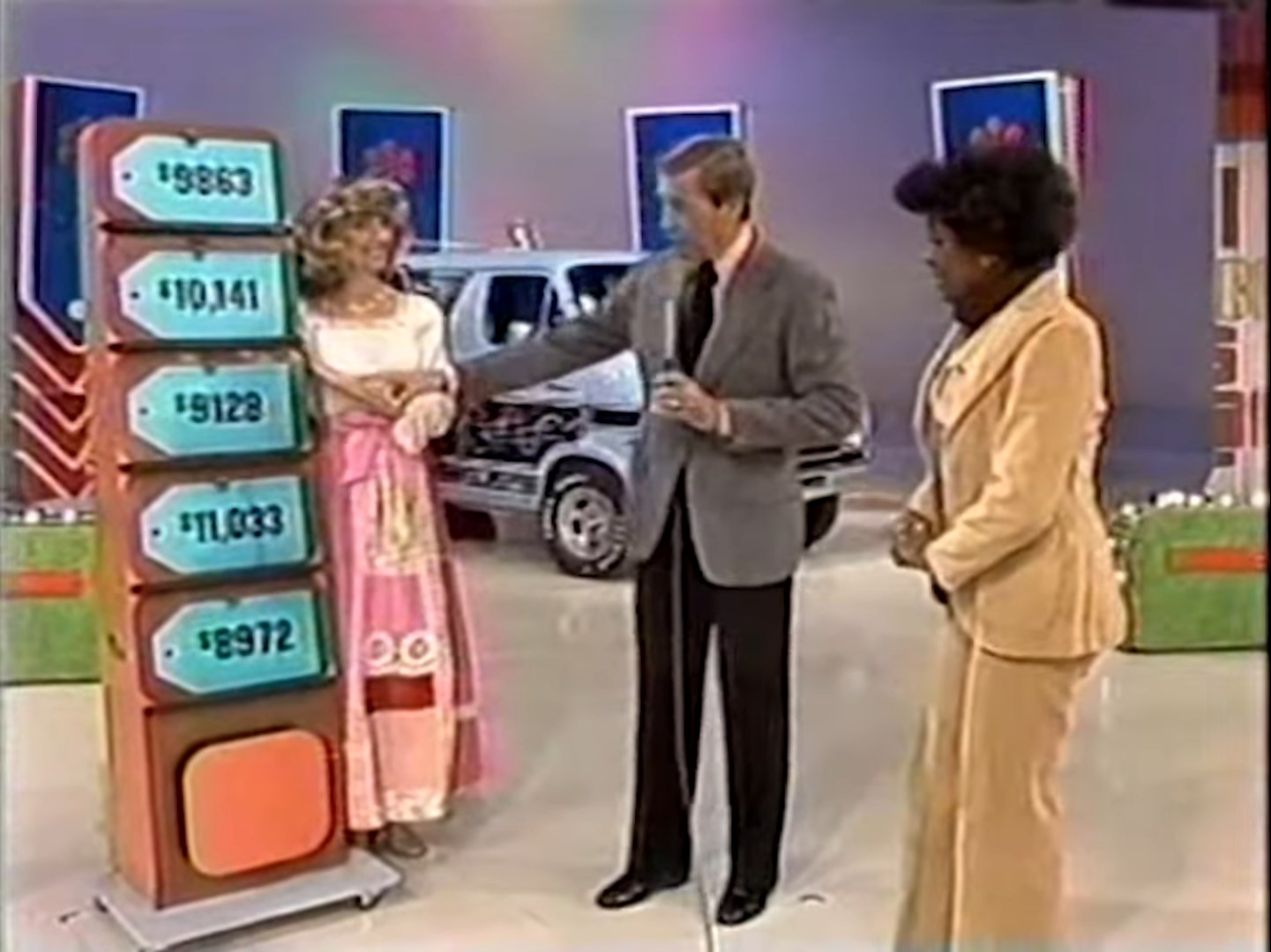 A game show setting featuring five colorful price tags displayed on a podium, with a contestant holding onto an arm of another person, while a host speaks next to them. A car is visible in the background.