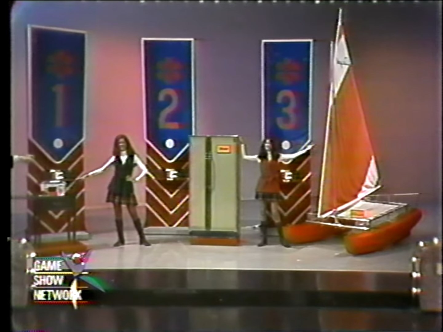 Two presenters showcasing three prize options labeled #1, #2, and #3 on a game show set, featuring a sewing machine, a refrigerator, and a catamaran.