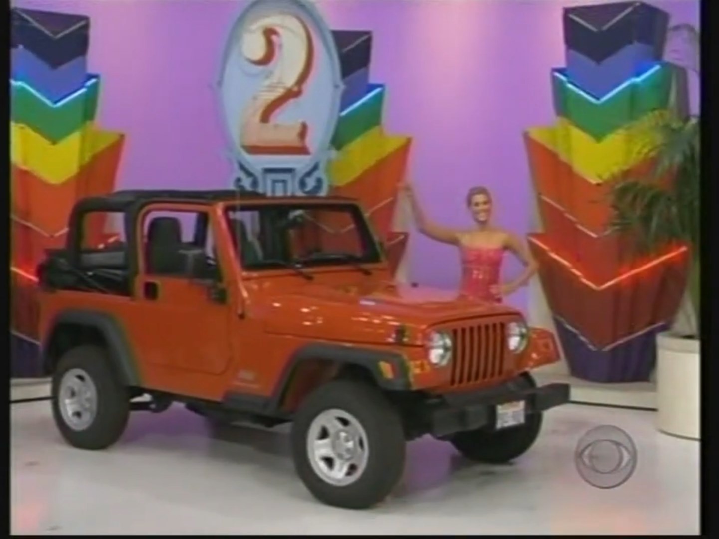 An orange Jeep Wrangler showcased on a game show set, with a smiling model standing next to it, highlighting the prize number two.
