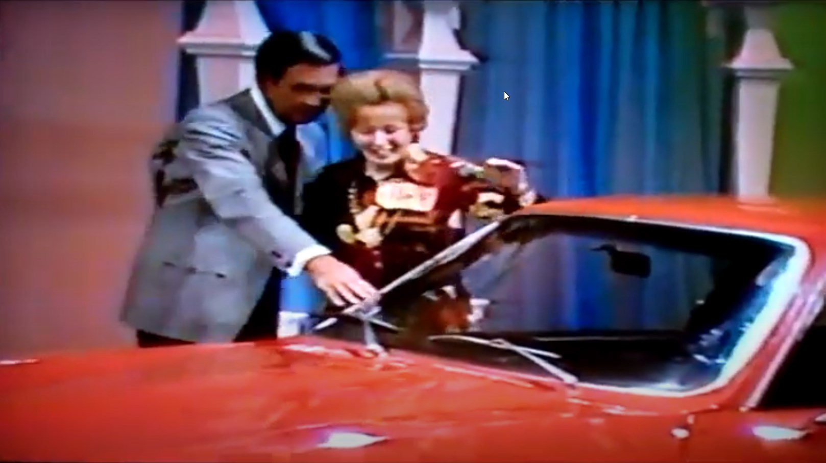 A man and a woman stand beside a bright red car, with the man pointing towards the car's windshield while the woman smiles excitedly.