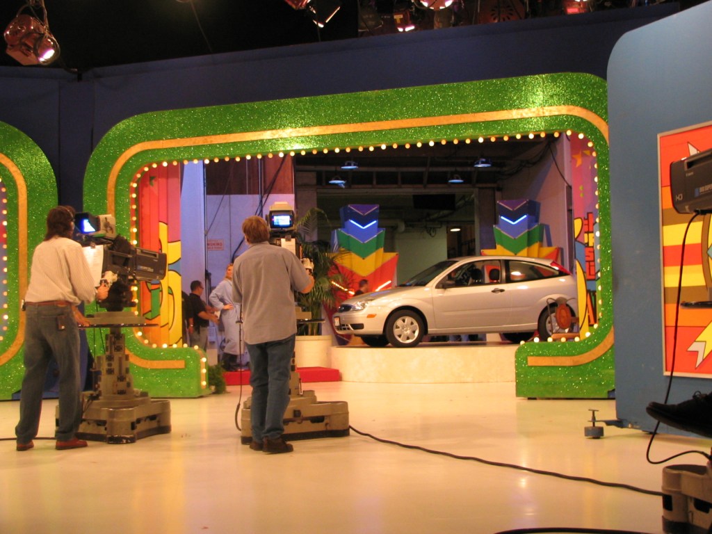 Behind the scenes view of a game show set with studio cameras and a silver car on stage, showcasing the vibrant colorful backdrop and lighting.