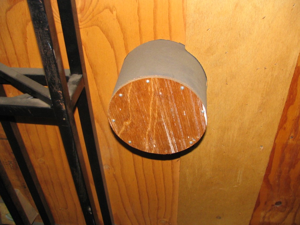 A wooden cylindrical object attached to a wall, featuring a metal casing and screws visible on its surface.