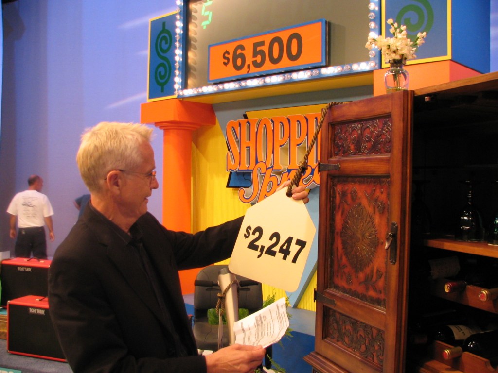 Roger Dobkowitz holds up a price tag of $2,247 during a game show segment in front of a colorful backdrop that features the words 'Shopping Spree'.