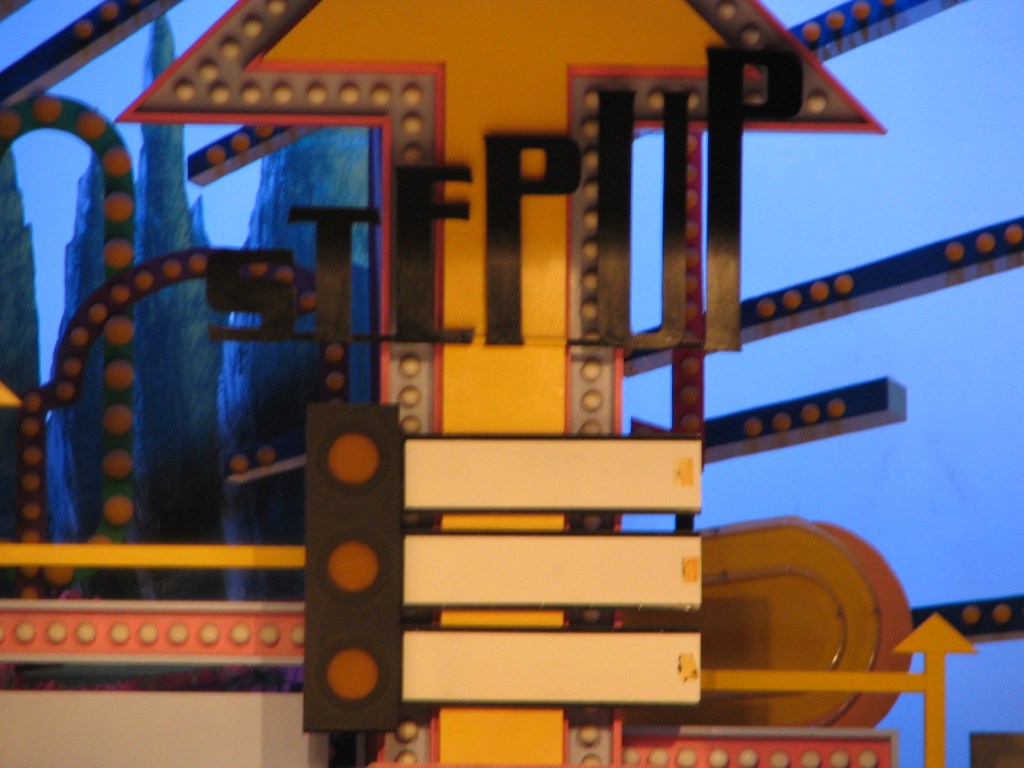 Close-up of a colorful stage prop featuring the word 'STEP UP' in bold letters, surrounded by decorative elements and lights.