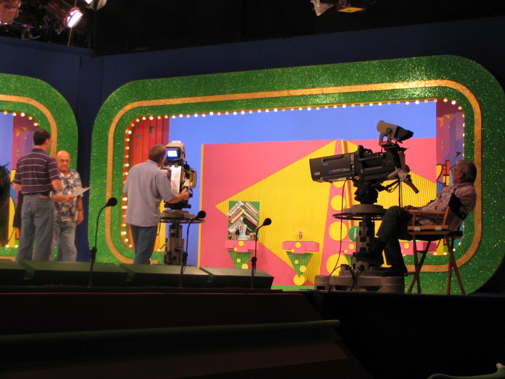 Behind-the-scenes of a game show set with crew members preparing, featuring colorful stage designs and camera equipment.