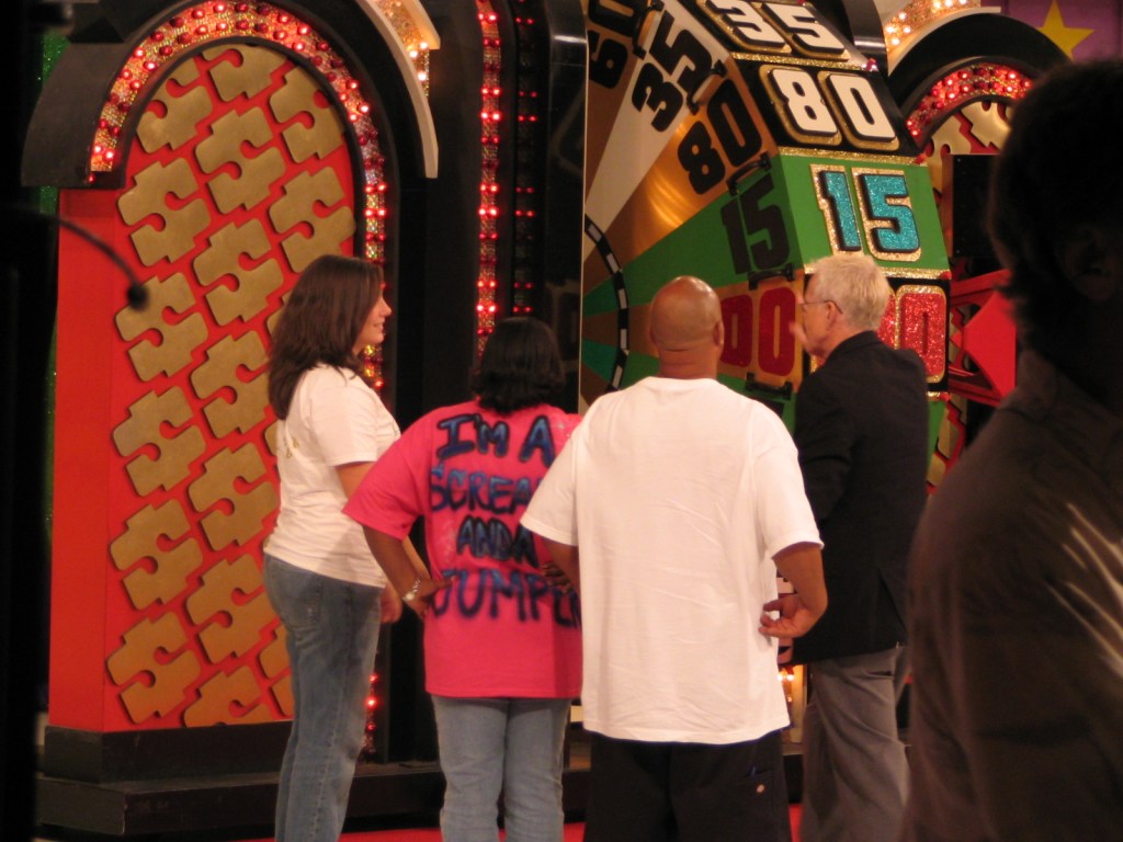 Three people standing in front of a large spinning wheel, with one person wearing a pink shirt that has the text 'I'm A Scream And I'm Jumping' visible on the back.